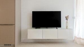 A modern living room with a flat-screen TV on a white cabinet, beige walls, and a window with sheer curtains.