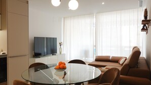 A modern living room with a glass dining table, brown leather sofa, and a flat-screen TV on a stand.