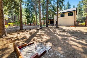 A forested area with a modern building, a fire pit, and a wooden bench.