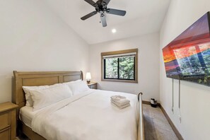 Ein Schlafzimmer mit Bett, Nachttisch, Deckenventilator, Fenster mit Blick auf Bäume und einem an der Wand montierten Fernseher.