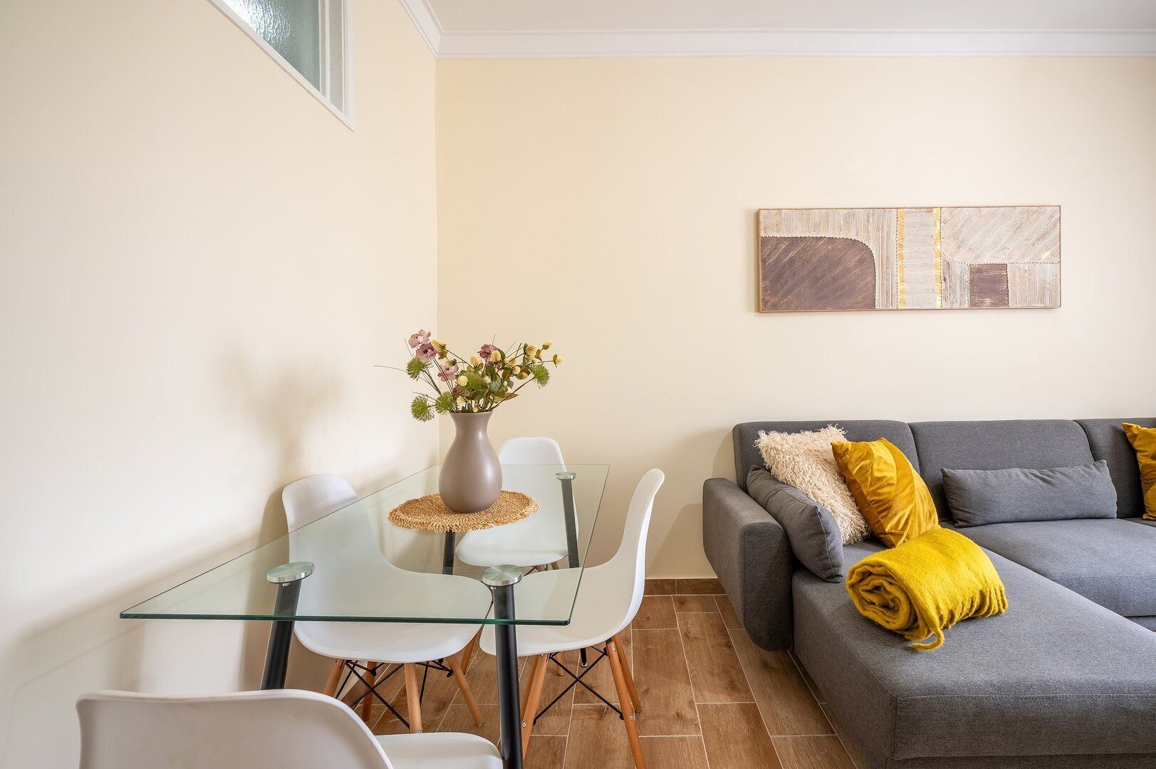 A modern living room with a glass dining table, white chairs, a grey sofa with yellow cushions, and a painting on the wall.