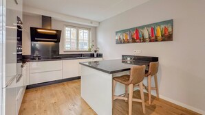 Une cuisine moderne avec des armoires blanches, un comptoir noir et un plancher en bois. On y trouve une fenêtre avec des stores, un tableau représentant des voiliers accroché au mur, et un petit îlot avec deux chaises.