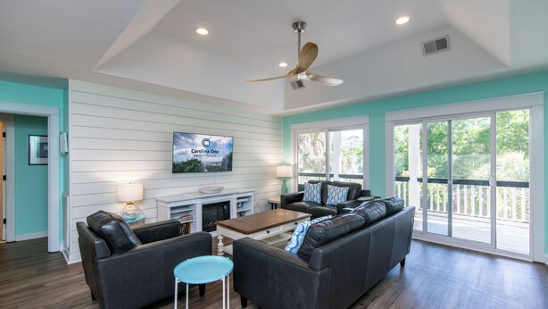 A spacious living room with a ceiling fan, a fireplace, a TV mounted on the wall, and large windows offering a view of the outdoors. - Knot in Kansas; Pet-Friendly, Sleeps 10, 1 Block to Beach, Firepit & Ping-Pong (Edisto Beach)