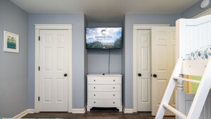A bedroom with a TV mounted above a dresser, two doors, a bunk bed, and a bed with a floral bedspread. - Knot in Kansas; Pet-Friendly, Sleeps 10, 1 Block to Beach, Firepit & Ping-Pong (Edisto Beach)
