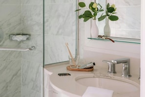 Un baño con pared de mármol, cabina de ducha con mampara de vidrio, lavabo blanco con grifo moderno y espejo sobre el lavabo.