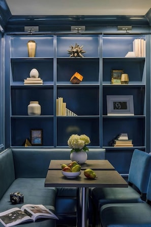 Una habitación con un sofá azul, una mesa con un florero y fruta, y una estantería con diversos objetos.