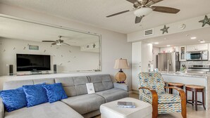 A modern living room with a grey sofa, blue pillows, a white ottoman, a patterned armchair, and a ceiling fan. - Pool Access: Beachfront Condo in Cape Canaveral! (Cape Canaveral)