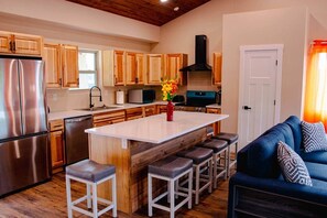 Una cocina moderna con armarios de madera, un refrigerador de acero inoxidable, una isla central con taburetes y un sofá azul con cojines estampados.