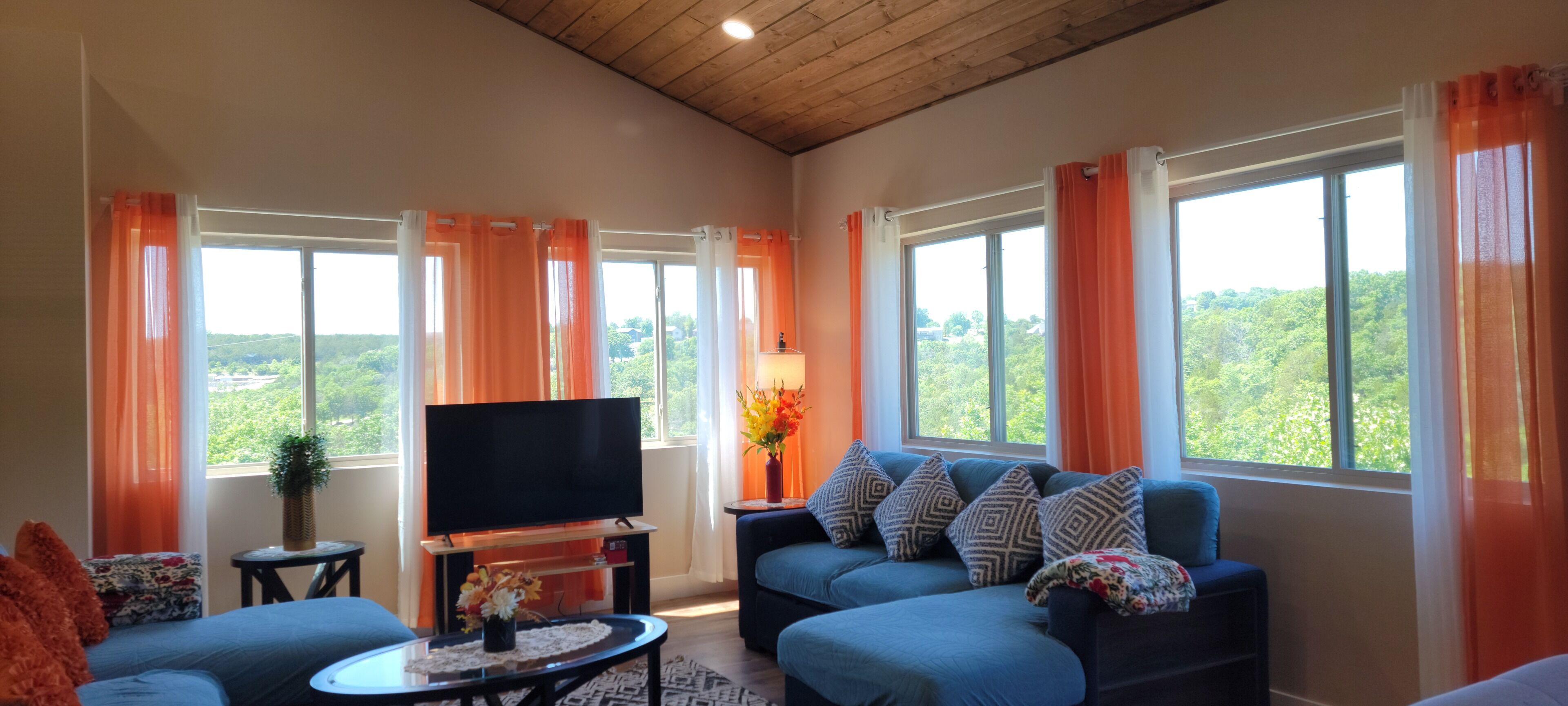 A living room with a TV, blue sofa, orange curtains, and a view of greenery outside.