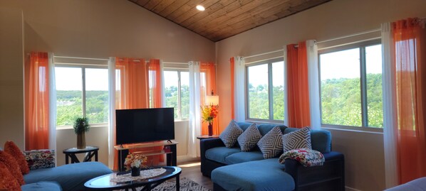 Un salón con televisor, sofá azul, cortinas naranjas y vistas al exterior con vegetación.