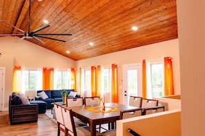 A spacious living area with a wooden ceiling, a dining table with chairs, a sofa, and a large window with curtains.