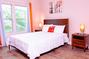A neatly made bed with white bedding and a red pillow, two bedside tables with lamps, a window with curtains, and a painting on the wall.
