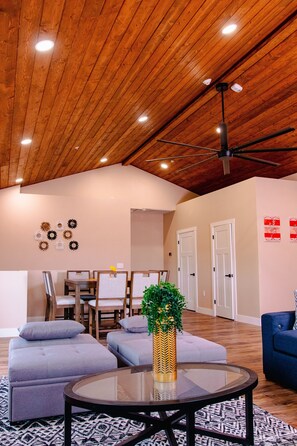 A spacious living area with a high wooden ceiling, a round glass table, grey sofas, and a decorative plant.