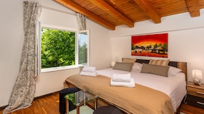 A bedroom with a large window, a bed with multiple pillows, a bedside table, and a painting on the wall.