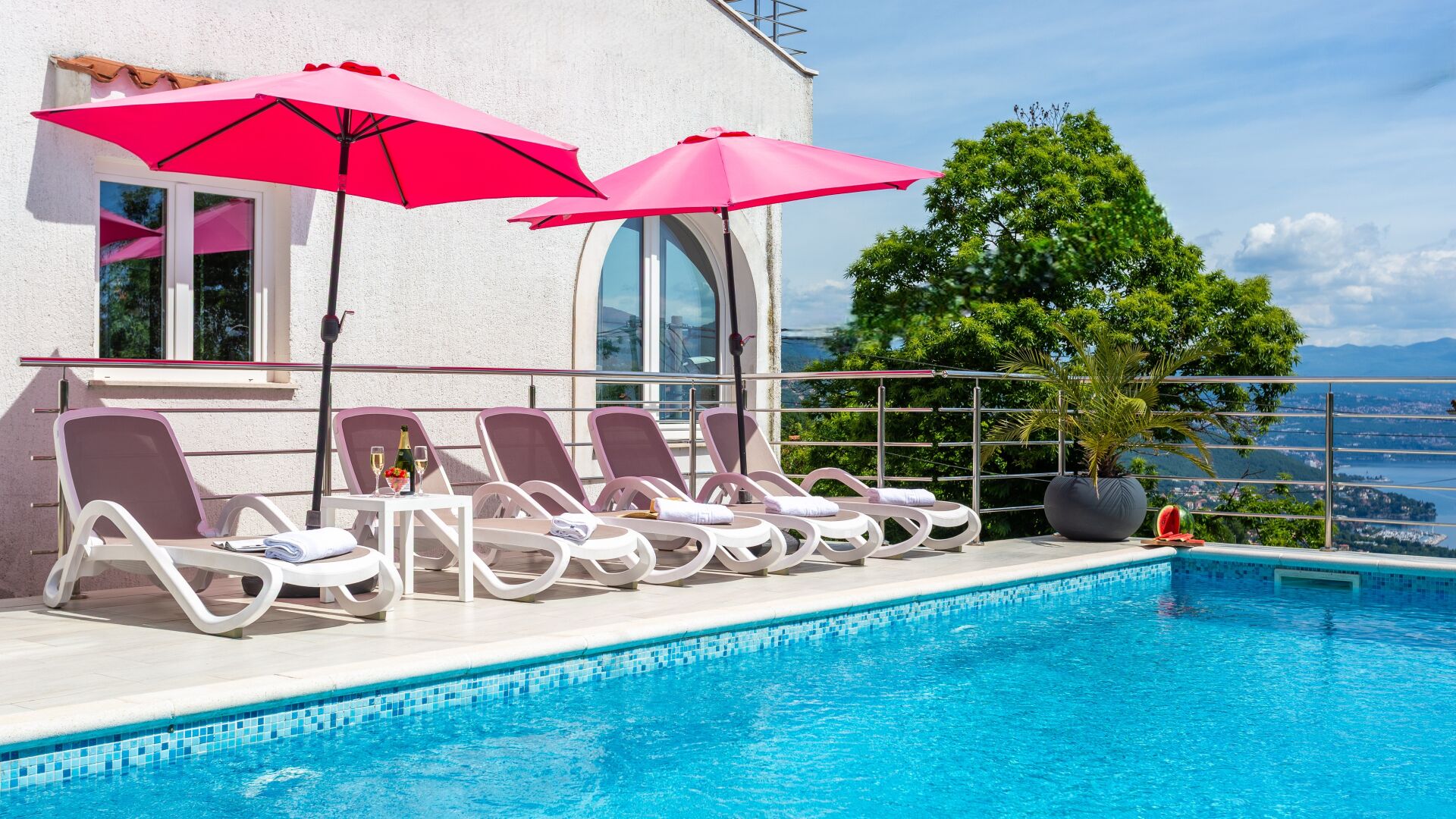 A poolside area with lounge chairs and umbrellas, a clear view of the sea, and a building with large windows.