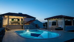 Une piscine avec une terrasse en pierre, un espace salon couvert et un escalier menant à un bâtiment doté d’un toit en tuiles.