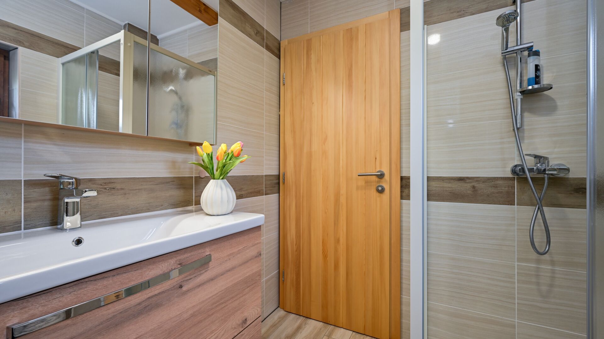 Une salle de bain avec une porte en bois, une douche, un lavabo agrémenté d’un vase de fleurs et un miroir.