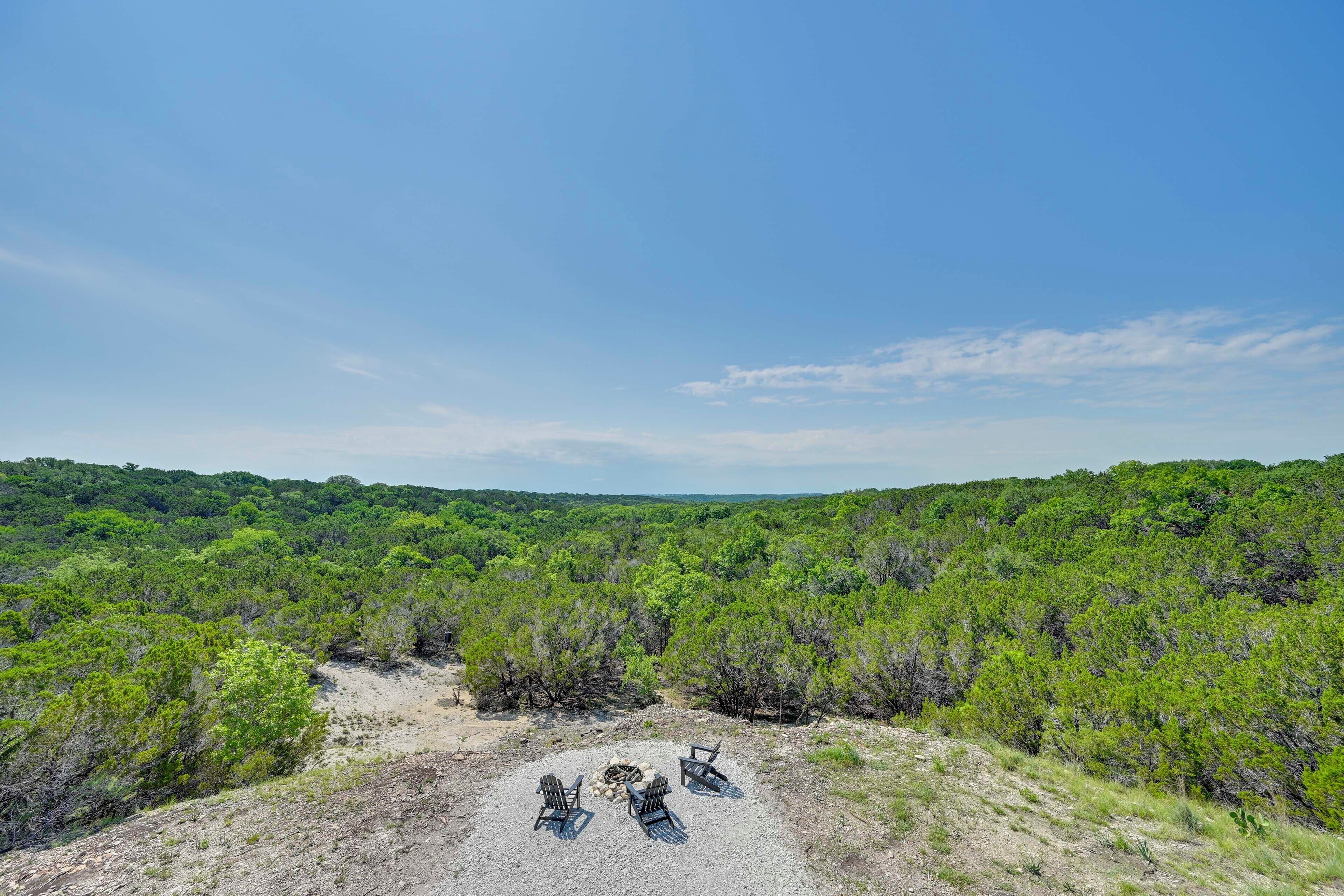 A natural landscape with a clear sky, dense green vegetation, and a group of Adirondack chairs around a fire pit.