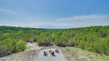 A natural landscape with a clear sky, dense green vegetation, and a group of Adirondack chairs around a fire pit.