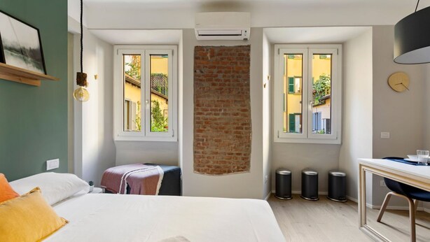 A hotel room with a bed, a desk, a chair, a window, and a brick wall. - GuestHost - Simplon & Garibaldi Comfy Studio (Milano)