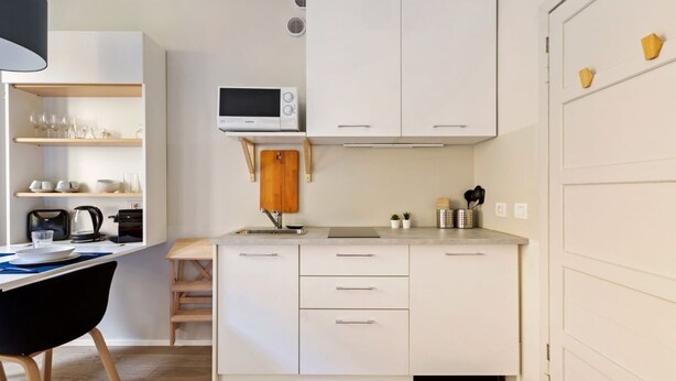 Cocina compacta con muebles blancos, microondas, lavamanos y comedor con una silla negra.