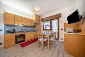 A modern kitchen with wooden cabinets, a dining table with chairs, a red rug, and a TV on the wall. - GuestHost - Lovely Mountain-Style Flat in Tarvisio (Tarvisio)