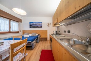 Une petite pièce comprenant un lit, une table, une chaise, un tapis rouge, une cuisine avec un évier et un tableau accroché au mur.