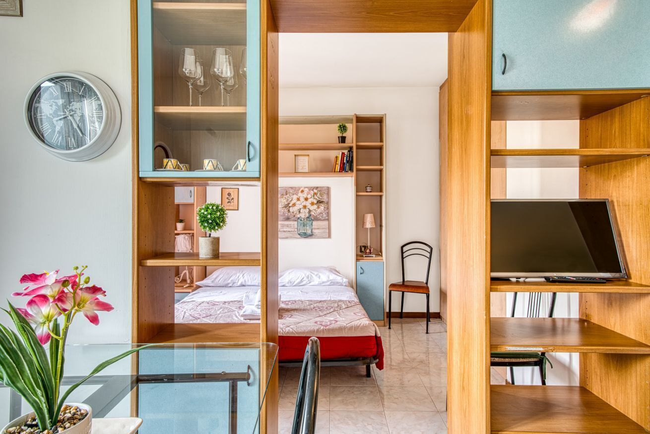 A room with a bed, a desk with a chair, a TV, and shelves with various items.