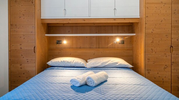 A single bed with a blue quilt, two white pillows, and a rolled towel on the bed. There is a wooden headboard with a shelf above the bed. - GuestHost - Cozy Apartment X3 in Tarvisio Center (Tarvisio)