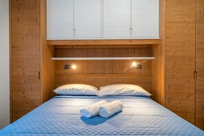 A single bed with a blue quilt, two white pillows, and a rolled towel on the bed. There is a wooden headboard with a shelf above the bed.