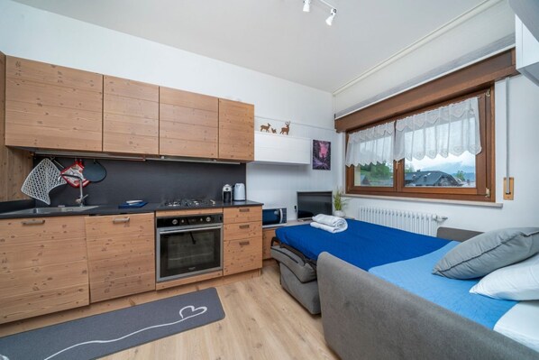 A compact room with a kitchenette, a bed with blue bedding, a grey sofa, and a window with a view of the outdoors.