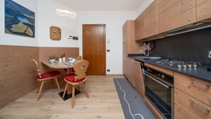 A compact kitchen with wooden cabinets, a dining table with chairs, a clock, and a painting on the wall. - GuestHost - Cozy Apartment X3 in Tarvisio Center (Tarvisio)