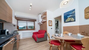 A compact kitchen with modern appliances, a dining area with wooden furniture, and a cozy seating area with a red blanket. - GuestHost - Cozy Apartment X3 in Tarvisio Center (Tarvisio)
