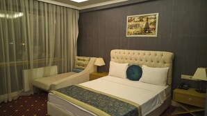 A hotel room with a large bed, two bedside tables, a window with sheer curtains, and a painting on the wall.