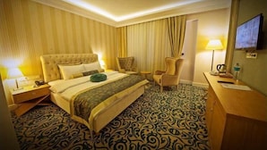 A hotel room with a large bed, a desk, a chair, a lamp, and a television on the wall. - Rabat Resort Hotel (Adiyaman)