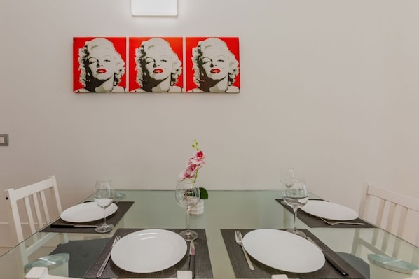 A dining table set with white plates, cutlery, and wine glasses, flanked by white chairs, with a red and white artwork above.