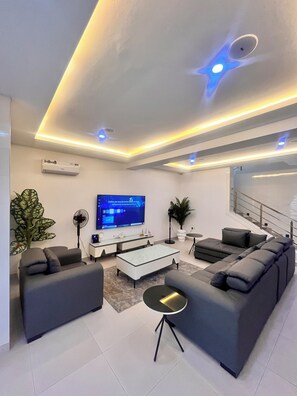 A modern living room with a flat-screen TV, grey sofas, a coffee table, and a staircase.