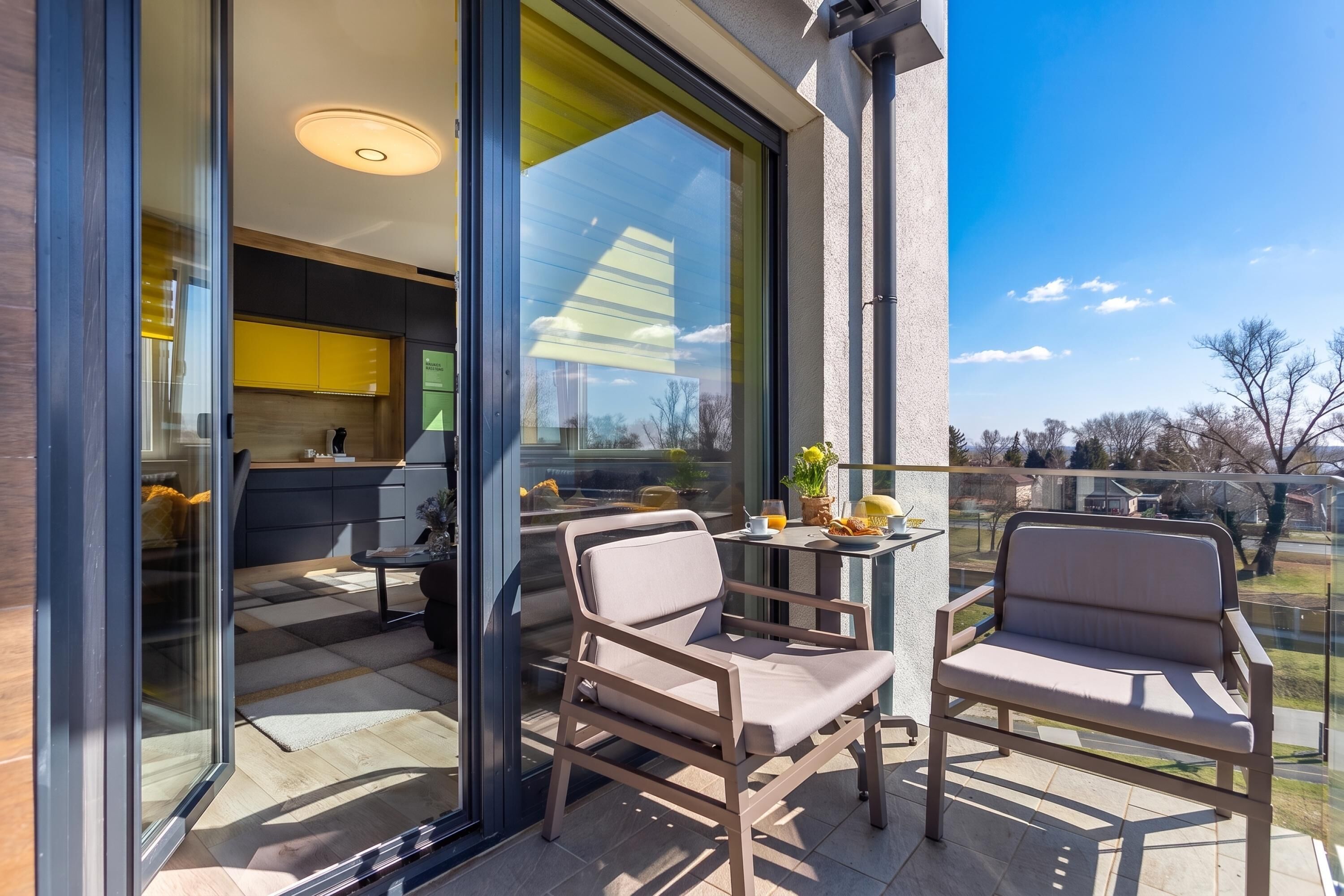 Un área de estar moderna al aire libre con dos sillas y una mesa, una puerta de vidrio que da a una cocina, y una vista despejada del cielo y el paisaje.