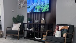 A modern living room with a flat-screen TV mounted on a wall, two dark sofas with patterned cushions, a wooden coffee table, and a decorative shelf with various items. - Tulum House in Austin Tx (Austin)