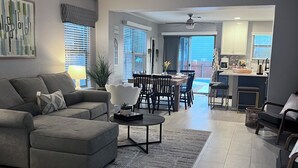 A modern living room with a gray sectional sofa, a coffee table, and a dining area with a table and chairs. There is a ceiling fan and a kitchen area visible in the background. - Tulum House in Austin Tx (Austin)