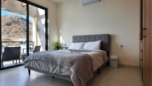 A bedroom with a bed, a nightstand, a potted plant, and a view of a mountainous landscape through large windows. - 2BD, 2BA Luxury Penthouse with Private Rooftop in Cabo.  LOTS OF BEACH GEARS! (Cabo San Lucas)