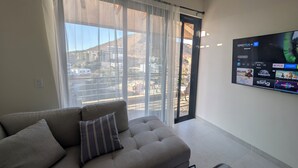 A living room with a sofa, a TV mounted on the wall displaying a streaming service interface, and a large window with sheer curtains. - 2BD, 2BA Luxury Penthouse with Private Rooftop in Cabo.  LOTS OF BEACH GEARS! (Cabo San Lucas)