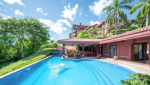 Una piscina con zona techada, rodeada de abundante vegetación y un edificio de fachada roja.