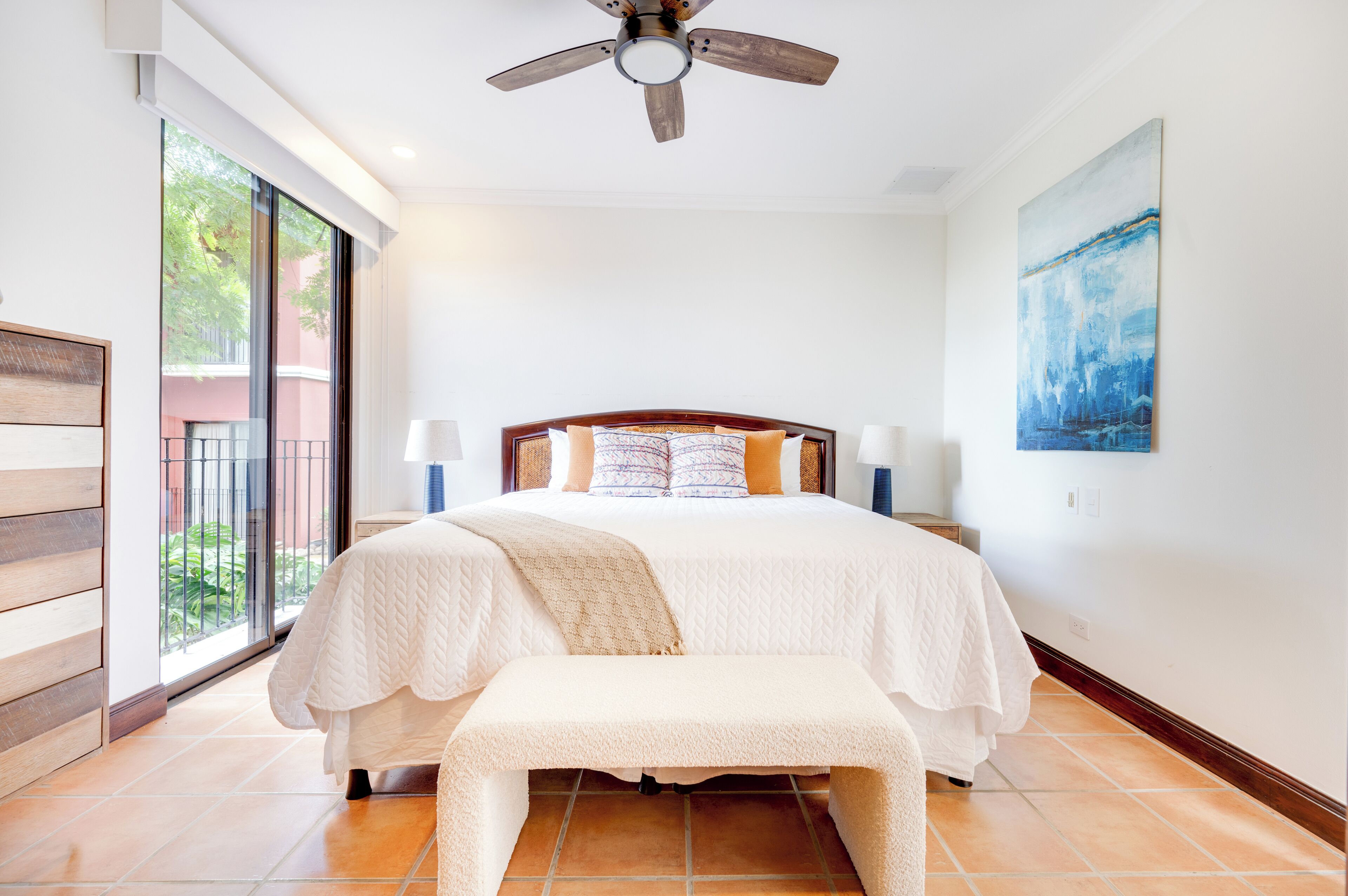 A bedroom with a bed, bedside lamps, a bench, a ceiling fan, and a large window.