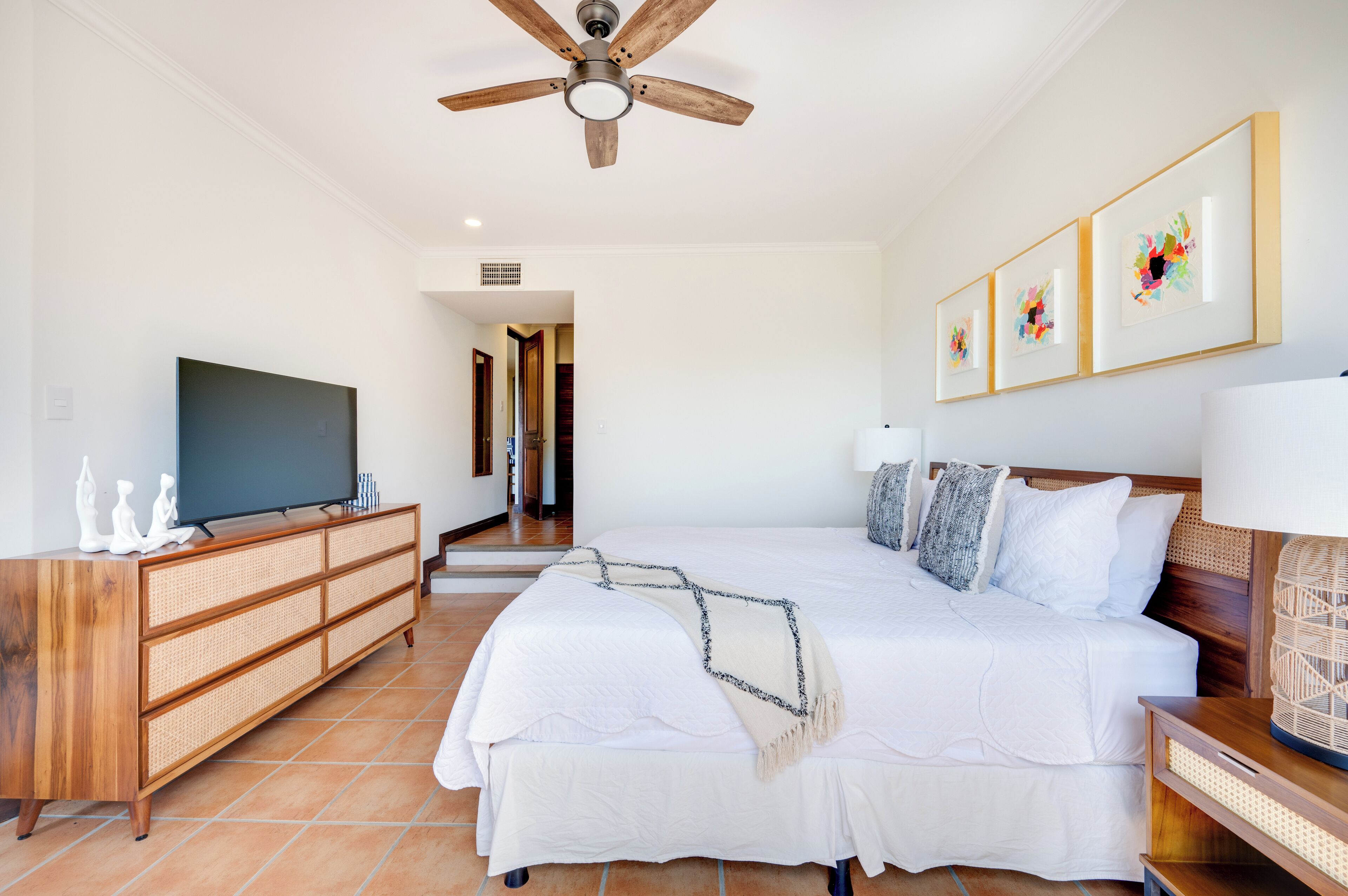 A spacious bedroom with a large bed, a wooden dresser, a ceiling fan, and a television.