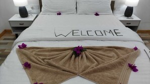Ein ordentlich bezogenes Bett mit einem 'WELCOME' Schild, zwei Nachttischlampen und ein braunes Handtuch mit violetten Blumen.
