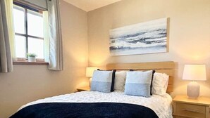 Una cama bien hecha con cubrecama blanco y azul marino, dos lámparas de noche, un cabecero de madera, una ventana con cortinas y un cuadro en la pared.