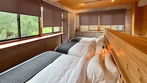 A bedroom with two beds, wooden headboards, large windows with blinds, and a stone wall.