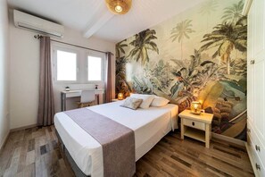 Quarto com cama, escrivaninha, cadeira, mesa de cabeceira com abajur e um mural tropical na parede.