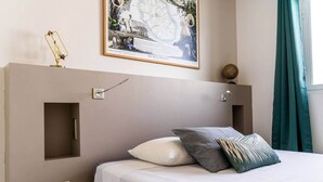 A bedroom with a bed, a nightstand, a wall-mounted lamp, a framed map, and a window with curtains.